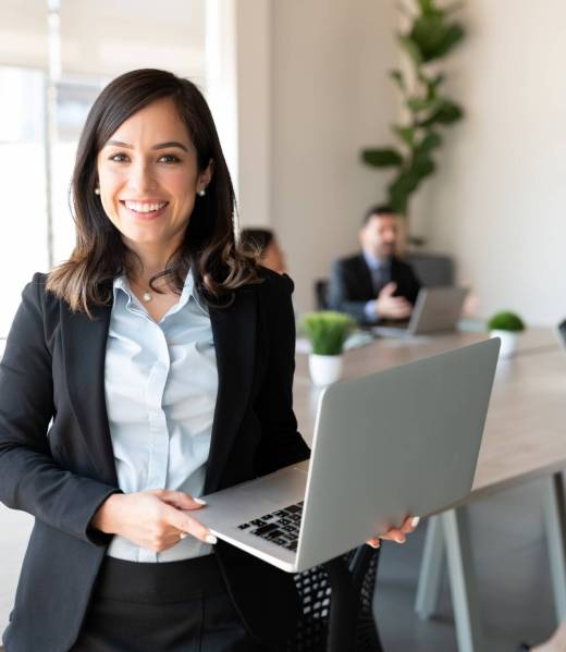Portrait of smiling female entrepreneur holding a laptop with team in background at office conference room