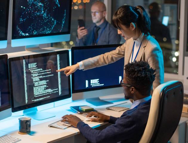 Young Asian businesswoman pointing at computer screen with decoded data while discussing it with African male colleague